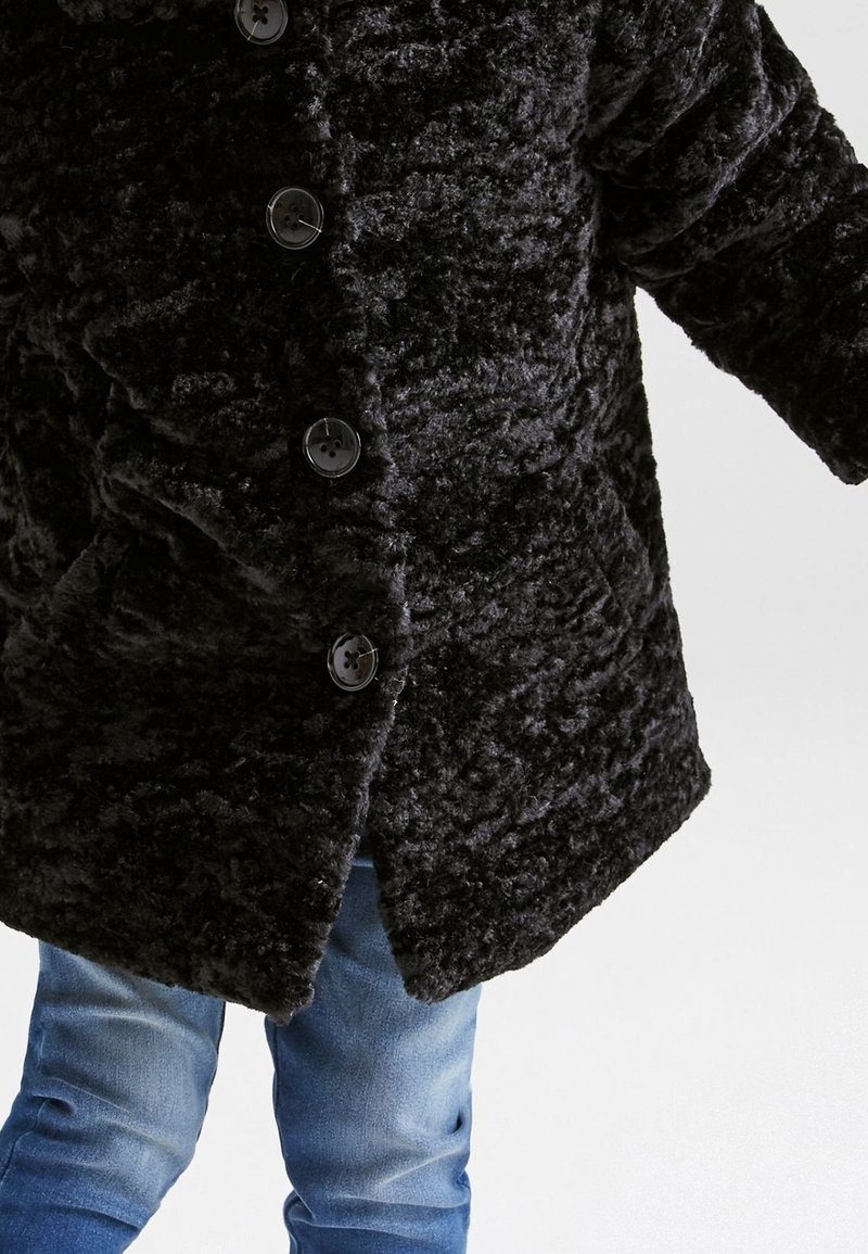 Enfant portant un manteau noir texturé boutonné avec un jean bleu sur un fond clair uni.