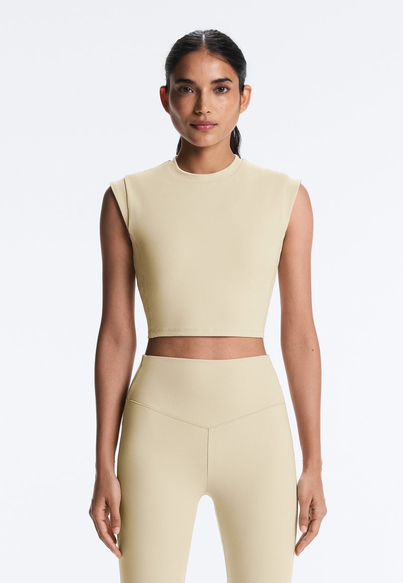 Tummatukkainen nainen ponnarilla, jolla on beige hihaton lyhyt toppi ja siihen sopivat korkeat trikoot, yksiväristä vaaleaa taustaa vasten.