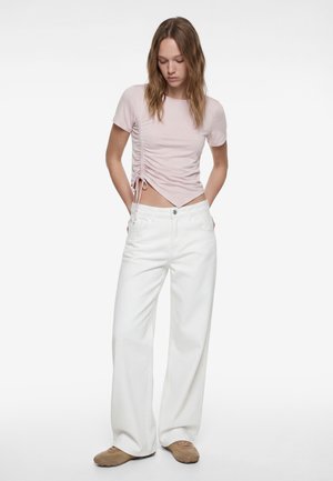 Frau trägt ein hellrosa gerafftes Kurzarm-Top, weiße Hosen mit weitem Bein und beigen Schuhen, steht mit den Händen in den Taschen und schaut nach unten.