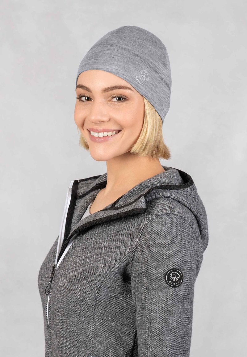 Bonnet gris avec une texture serrée et lisse, arborant un logo discret sur le côté. Porté avec un sweat à capuche zippé gris. Design doux et décontracté.