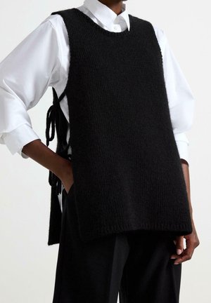 Schwarzer, gestrickter, ärmelloser Pullover mit rundem Ausschnitt, offene Seiten mit Stoff gebunden, getragen über einem weißen Hemd mit Kragen, kombiniert mit dunkler Hose.