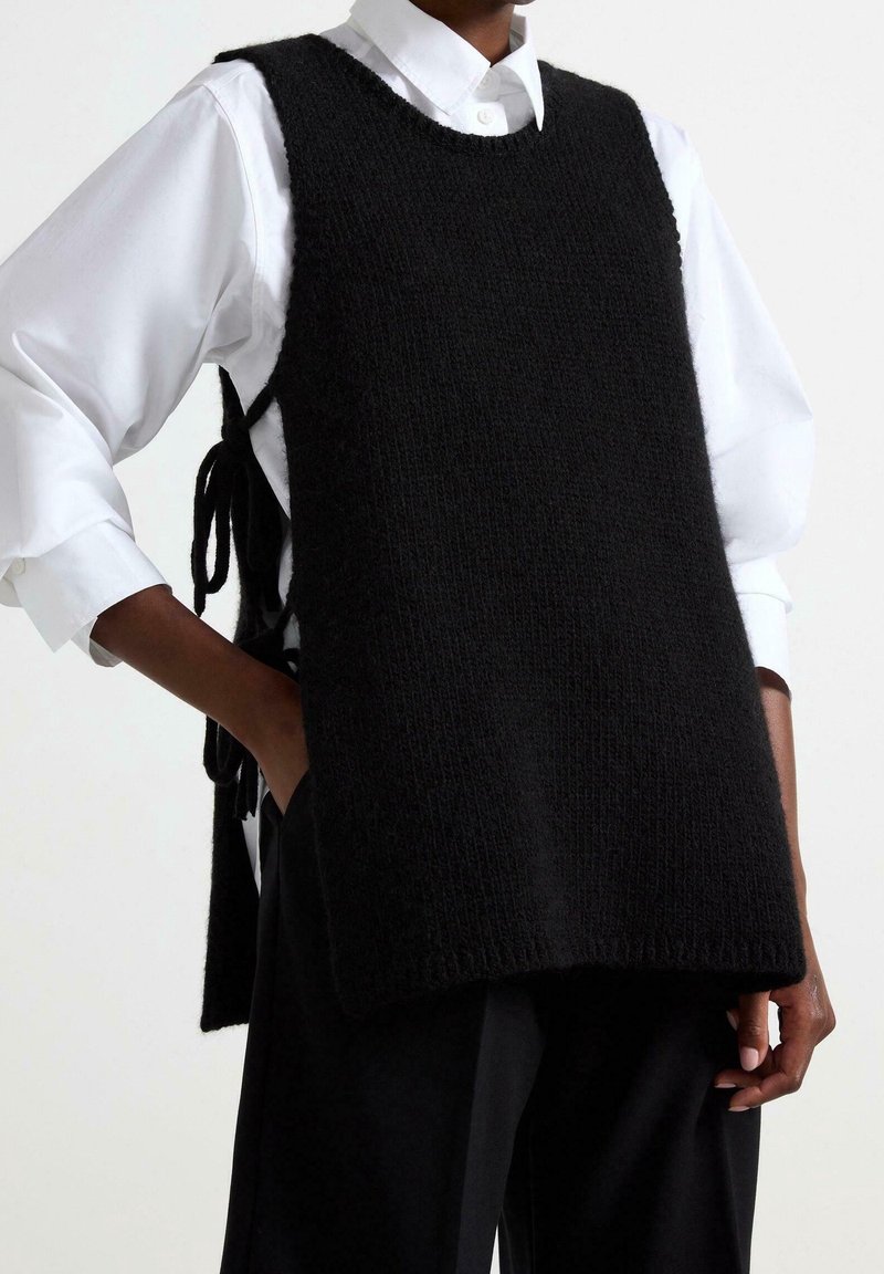 Pull sans manches en maille noire avec un col rond, côtés ouverts noués avec du tissu, porté sur une chemise blanche à col, associé à un pantalon foncé.