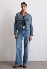 Veste en denim bleu clair avec col beige, dotée de deux poches poitrine et d'une fermeture à boutons. Associée à un jean bleu clair taille haute.