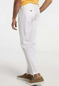 Pantalon blanc avec une coupe ajustée, comportant une poche arrière avec un bouton noir. Porté avec des chaussures à enfiler marron clair.