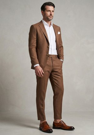 Mann mit kurzem dunklem Haar und Bart, trägt einen braunen Anzug, weißes Hemd, weißes Einstecktuch und braune Lederschuhe, steht vor einem grauen Hintergrund.