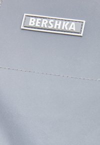 Šedá tkanina s hladkým povrchem, s obdélníkovou stříbrnou jmenovkou se značkou "BERSHKA" v bílých písmenech a jemným prošíváním podél okrajů.