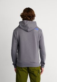 Sudadera gris con capucha ajustable, puños y dobladillo acanalados, que presenta un logotipo azul en la parte superior de la espalda. Llevada con pantalones verdes.