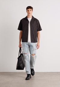 Camisa negra de manga corta con un patrón de logotipos repetidos, combinada con jeans rotos de color azul claro y zapatos de cuero negros, llevando una bolsa tote negra.