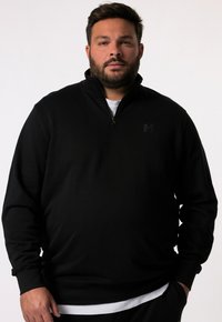 Felpa nera con zip e texture a coste, caratterizzata da un colletto alto e un logo discreto sul petto, indossata sopra una camicia bianca.