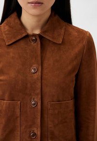 Bruine suède jas met een kraag, voorzien van een knopenrij en twee grote zakken. De textuur is glad met een zachte uitstraling.