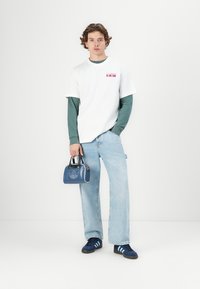 Hvit t-skjorte lagdelt over en grønn langermet skjorte, lyseblå jeans og marineblå sneakers. Holder en liten blå håndveske med logodetaljer.