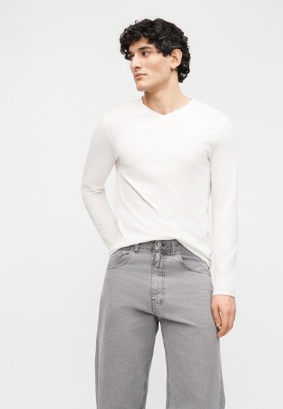 Νεαρός με σγουρά μαλλιά, φορώντας λευκό πουκάμισο μακρυμάνικο και ανοιχτόχρωμα γκρι τζιν, στέκεται με το ένα χέρι στην τσέπη του.