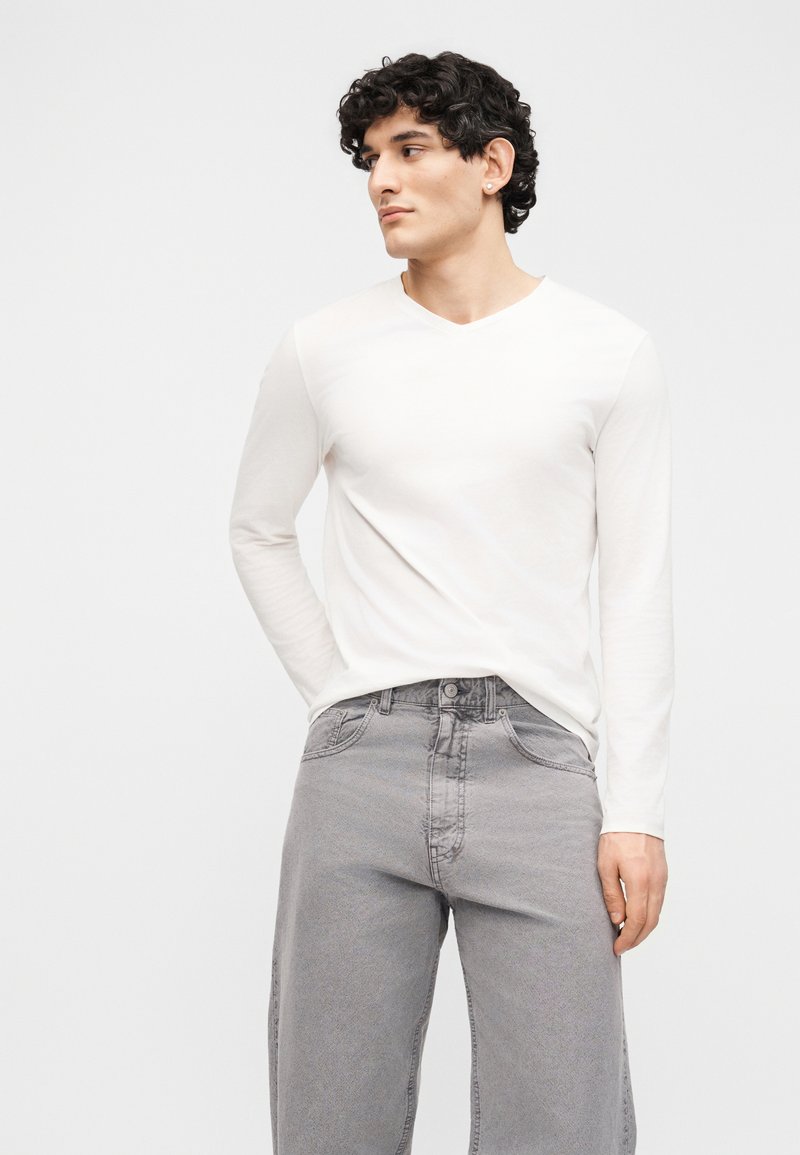 Jeune homme aux cheveux bouclés portant une chemise blanche à manches longues et un jean gris clair, debout avec une main dans sa poche.