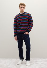 Multicolored striped sweater with a crew neck, featuring red, green, blue, and black stripes; paired with dark jeans and white sneakers.