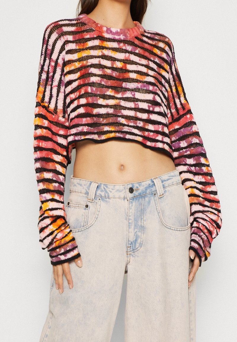 Nainen, joka on pukeutunut lyhytmalliseen, moniväriseen raidalliseen neuleeseen, jossa on pitkät hihat, sekä korkeavyötäröisiin vaaleansinisiin leveisiin farkkuihin.