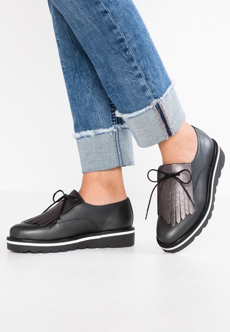 Chaussures en cuir noir avec un design à franges et des lacets noirs, dotées d'une semelle blanche épaisse et d'accents texturés, assorties à un jean bleu retroussé.
