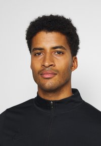 Zwarte Nike zip-up jack met een hoge kraag, een gladde textuur en een subtiele logo aan de linkerkant. Model heeft kort, krullend haar.