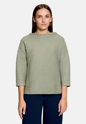 Vrouw met stijl bruin haar, gekleed in een gestructureerde lichtgroene top met lange mouwen en donkere broek, staand tegen een effen lichte achtergrond.