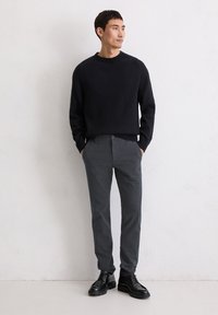 Schwarzer gerippter Pullover mit langen Ärmeln, kombiniert mit dunkelgrauen Slim-Fit-Hosen und schwarzen Schnürschuhen. Einfarbiger Hintergrund.