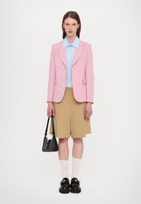 Blazer rosa chiaro sopra una camicia azzurra, abbinato a pantaloni corti beige. Borsa nera, calzini bianchi e scarpe nere dal design robusto.