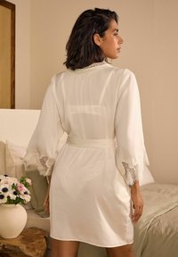 Robe en satin crème avec des manches cloche et des accents en dentelle au niveau des poignets. Liée à la taille, avec une délicate broderie au niveau du col.