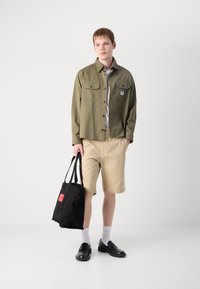 Oliven grønn knapp-up skjorte, beige shorts, svarte slip-on sko, hvite sokker, holder en svart tote bag med rød aksent. Enkel, uformell antrekk.