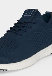 Baskets de sport bleu foncé avec tige tricotée, lacets et semelle blanche texturée, affichant le logo de la marque sur la languette.