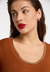 Brun strikket sweater med en bred scoop halsudskæring, der har en guldkæde accent. Modellen har langt hår, gyldne hoops ørehængere og kraftige røde læber.