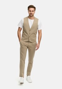 Chaleco beige con cinco botones, combinado con pantalones a juego. Una camiseta blanca lisa debajo y zapatillas blancas completan el conjunto.