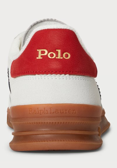 Sapatilha branca com aba de calcanhar em pele vermelha, apresentando "Polo" em dourado. Sola de borracha em tom bege com "Ralph Lauren" em relevo. Parte superior texturizada acrescenta detalhe.