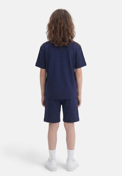 Otrok z ramensko dolžino kodrastih las stoji obrnjen stran, oblečen v mornarsko modro majico in kratke hlače ter bele nogavice in superge.