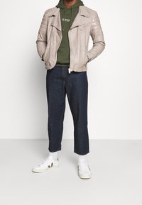 Giacca da motociclista in pelle beige con spalle trapuntate sopra una felpa con cappuccio verde oliva. Jeans blu scuro tagliati e sneakers bianche con dettagli grigi.
