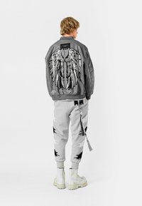 Chaqueta bomber gris con grandes alas de ángel de lentejuelas en la espalda, combinada con pantalones jogger grises con patrones en blanco y negro, y botas de combate blancas.