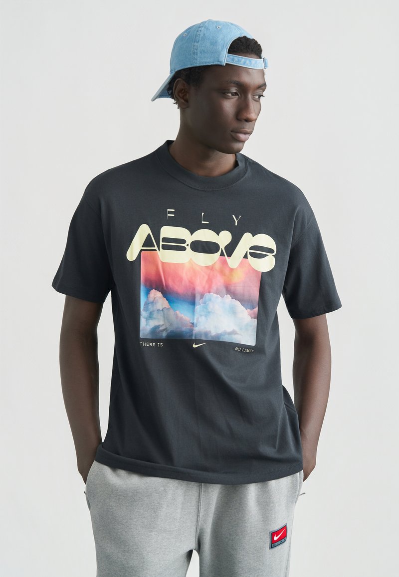 Μαύρο βαμβακερό μπλουζάκι με γραφικό τύπωμα νεφών σε παστέλ χρώματα. Το κείμενο γράφει «FLY ABOVE» με έντονα κίτρινα γράμματα. Κοντά μανίκια.