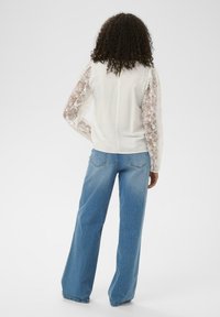 Haut blanc avec manches longues en dentelle florale transparente et un petit trou dans le dos. Associé à un jean bleu clair taille haute à jambes larges.