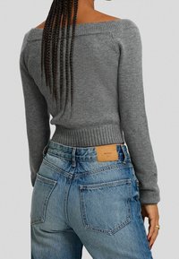 Maglione grigio off-the-shoulder con orlo a coste abbinato a jeans in denim blu a vita alta con una fodera in pelle beige e dettagli di cucitura visibili.