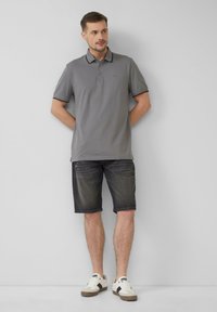 Grijze poloshirt met zwarte details, gecombineerd met donkere denim shorts. Het model draagt witte sneakers met grijze accenten en staat tegen een effen achtergrond.
