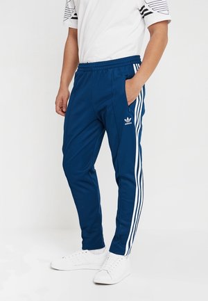 Osoba ubrana w niebieskie spodnie dresowe Adidas z białymi paskami i logo, w połączeniu z białymi tenisówkami i białą koszulką z krótkim rękawem.