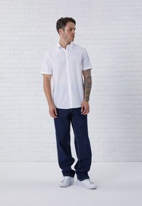 Camisa blanca de manga corta con botones, vaqueros oscuros de corte ancho y zapatillas blancas. Con un fondo de ladrillo blanco.