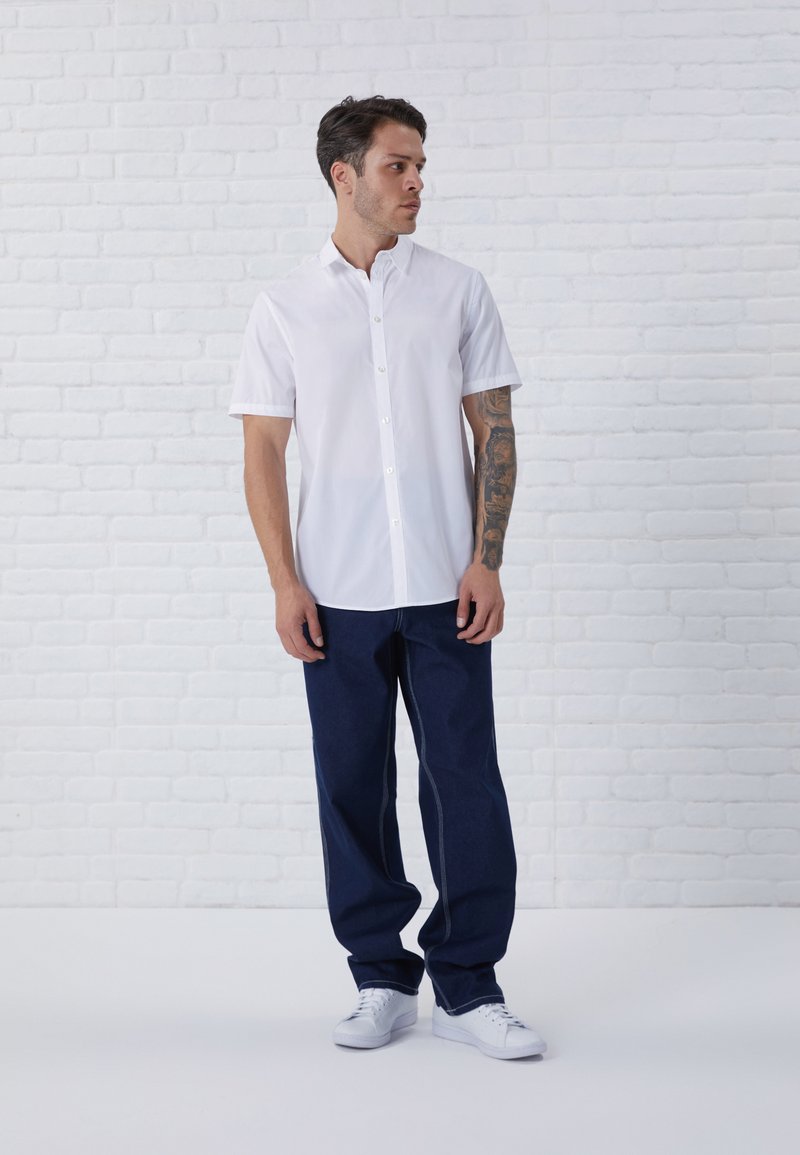 Camisa blanca de manga corta con botones, vaqueros oscuros de corte ancho y zapatillas blancas. Con un fondo de ladrillo blanco.