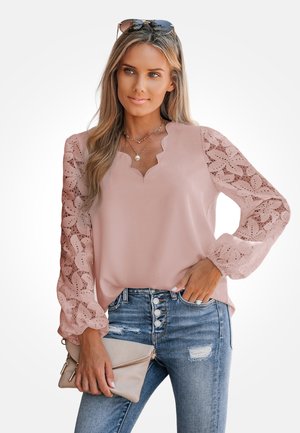 Blusa rosa con un escote festoneado y mangas transparentes de estilo encaje con patrones de hojas. Combinada con vaqueros azules desgastados y un bolso de mano.