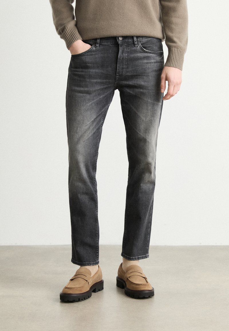 Jeans din denim de culoare gri închis cu un finisaj estompat, având un design clasic cu cinci buzunare și o croială slim, purtate cu mocasini de culoare maro deschis.