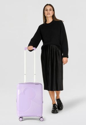 Valigia rigida viola con motivi a linee curve, manico retrattile e ruote nere, abbinata a un maglione nero e una gonna plissettata.