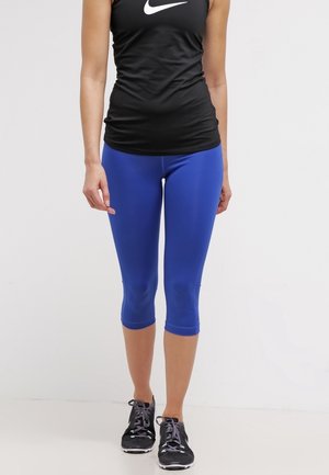 Femme portant un débardeur noir Nike, des leggings courts bleu vif et des chaussures de course noires, debout devant un fond blanc uni.