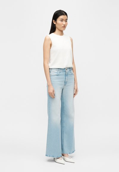 Γυναίκα με μακριά μαύρα μαλλιά, φορώντας λευκό αμάνικο τοπ, ανοιχτό μπλε τζιν παντελόνι με φαρδιά μπατζάκια και λευκά μυτερά τακούνια, στέκεται μπροστά σε απλό φόντο.