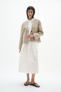 Frau mit glattem, dunklem Haar, die einen beigen Blazer, ein weißes, geknöpftes Hemd, einen langen weißen Rock mit wellenförmigem Saum und schwarze Flip-Flop-Sandalen trägt.