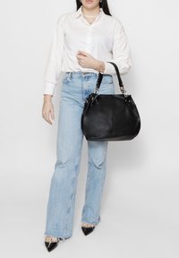 Borsa in pelle nera con forma arrotondata e metalleria argentata. Indossata da una persona con camicia bianca e jeans azzurri con orli sfrangiati.