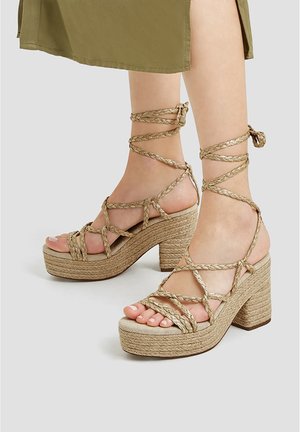Sandales compensées en or tressé avec un talon en jute texturé, des brides croisées à la cheville et des orteils ouverts. Couleur : beige clair.