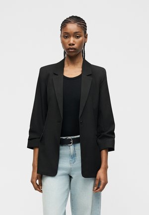 Junge schwarze Frau mit geflochtenem Haar, trägt einen schwarzen Blazer über einem schwarzen Oberteil und helle blaue Jeans mit einem schwarzen Gürtel, steht vor neutralem Hintergrund.