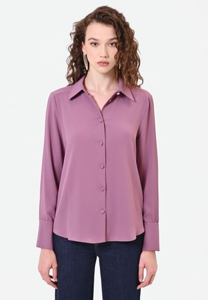 Donna con capelli ricci che indossa una camicetta lilla a maniche lunghe con bottoni e jeans blu scuro, in piedi davanti a uno sfondo semplice.